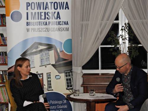 Autorka Katarzyna Pełczyńska-Nałęcz oraz prowadzący siedzący przy stoliku podczas rozmowy na spotkaniu w bibliotece.